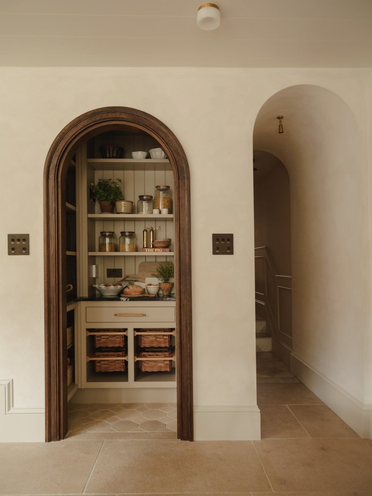 Arched wooden pantry doorway with white shelving and woven baskets inside a cream room.