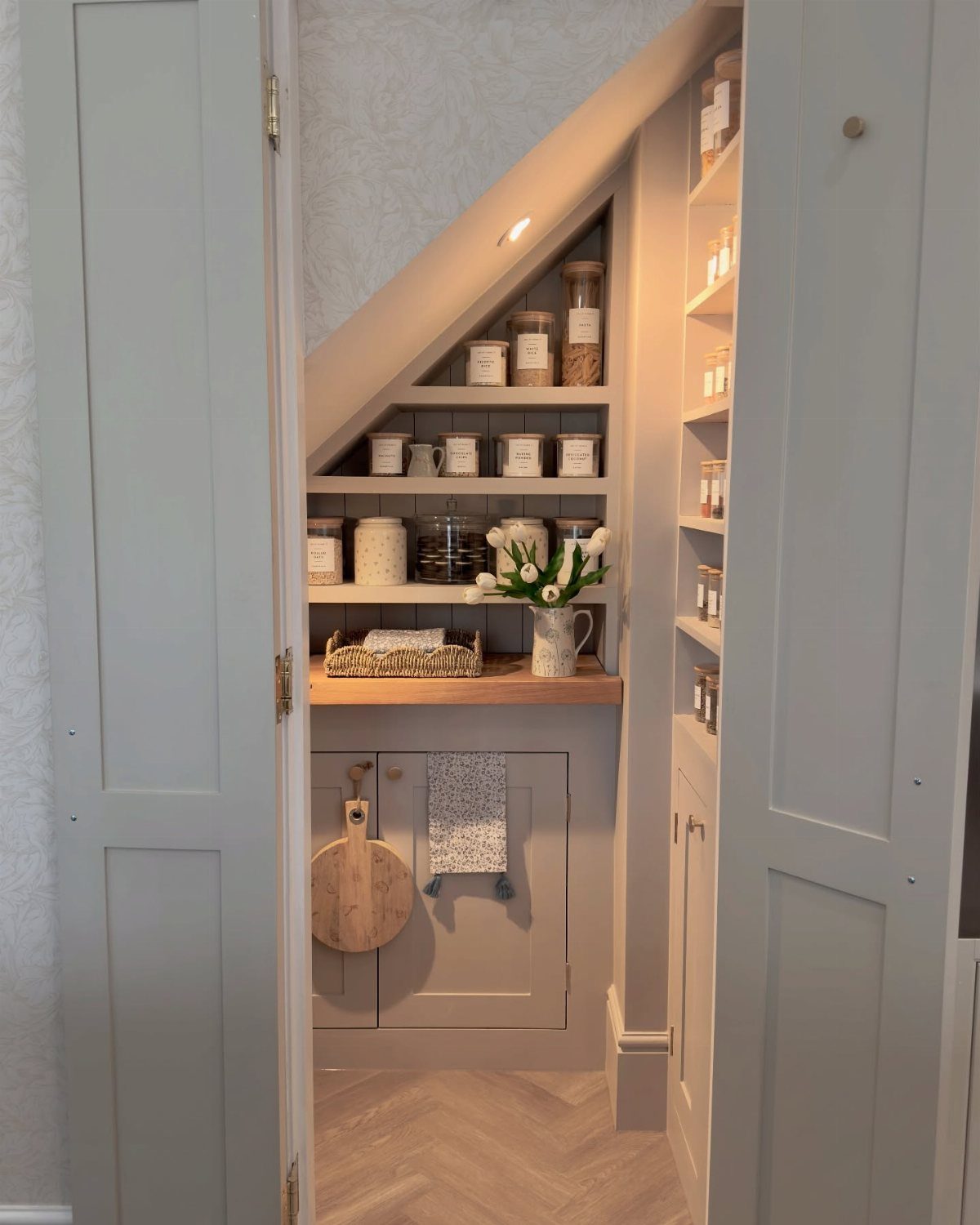 Under-stairs pantry with light grey cabinets and open shelves holding decorative food jars.