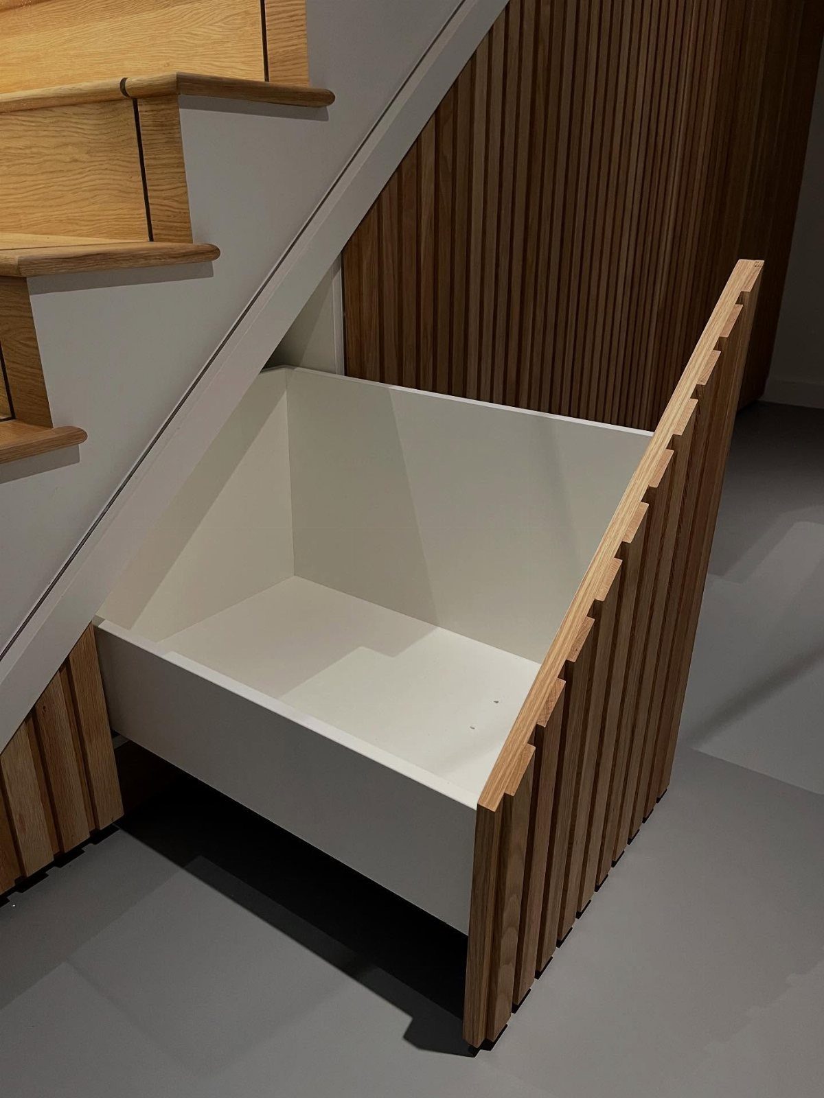 White pull-out storage drawer built into the space under a wooden staircase.
