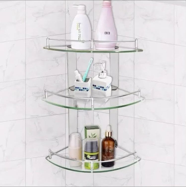 Three transparent glass corner shelves holding toiletries on a white tiled bathroom wall.