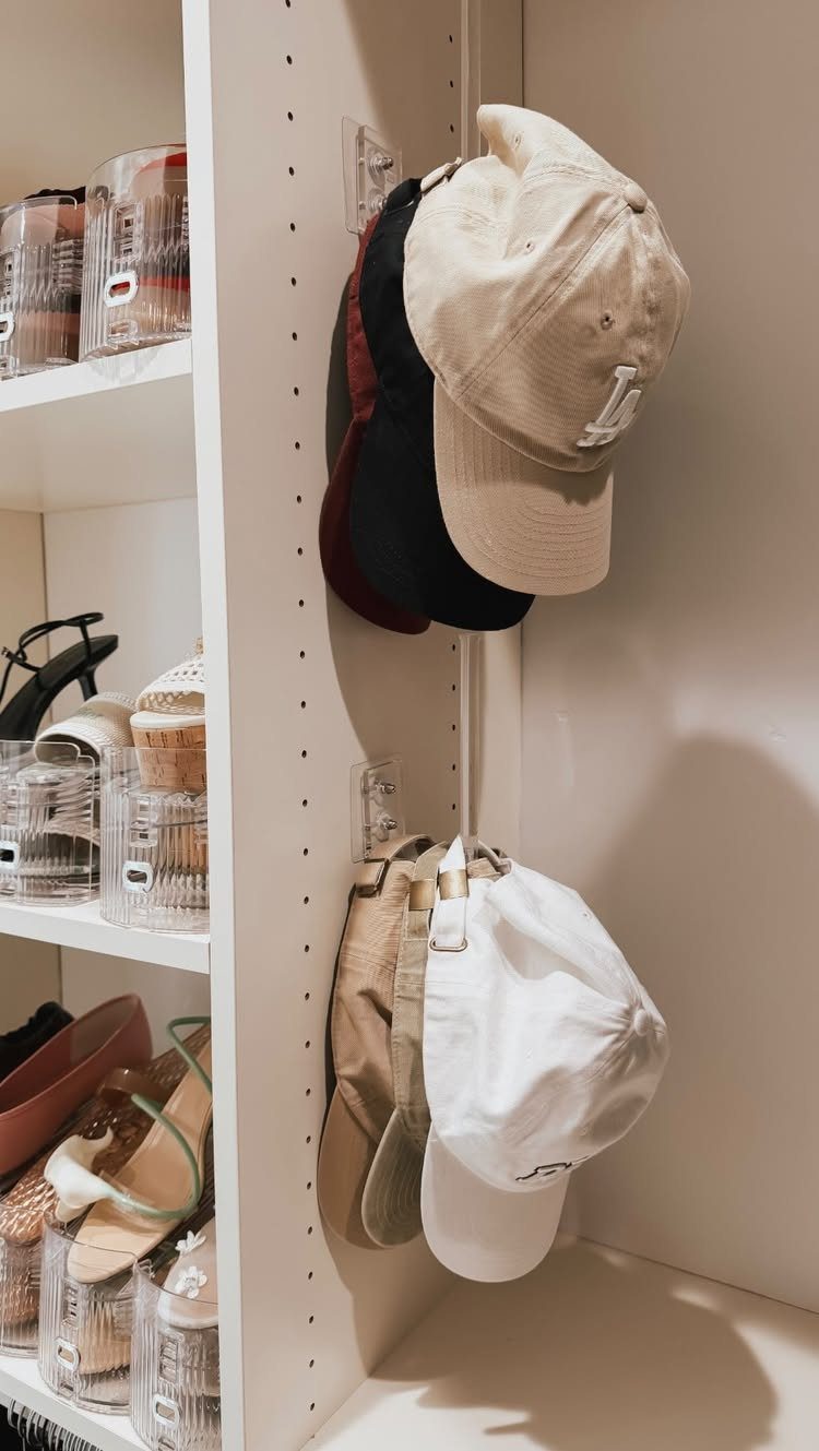 Baseball caps hanging from hooks inside a white closet with shoe storage shelves.
