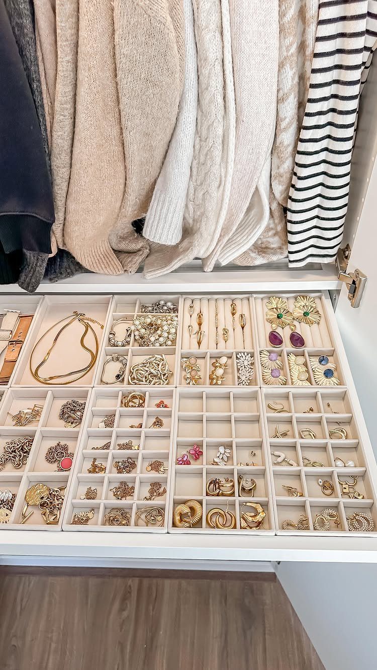 Jewelry organized in beige velvet trays inside a white closet drawer below hanging clothes.