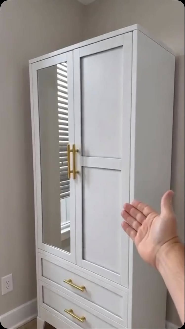 White freestanding wardrobe with a mirrored door and gold handles against a grey wall.