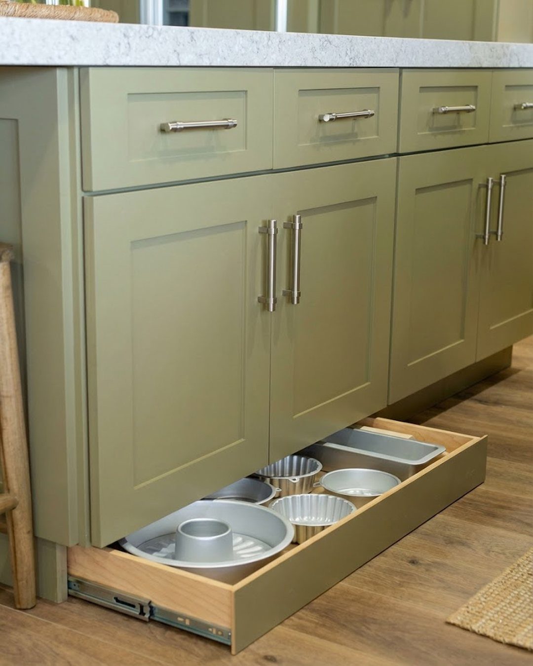 Toe-kick drawer under olive green kitchen cabinets holding various metal baking pans.