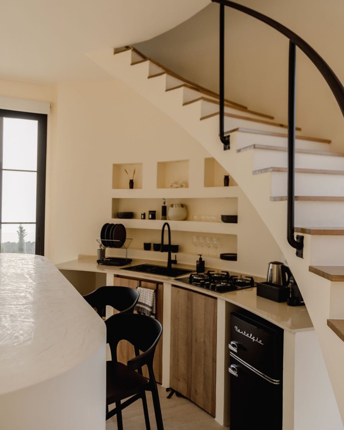 Kitchenette with recessed open shelving built into the wall under a curved staircase.