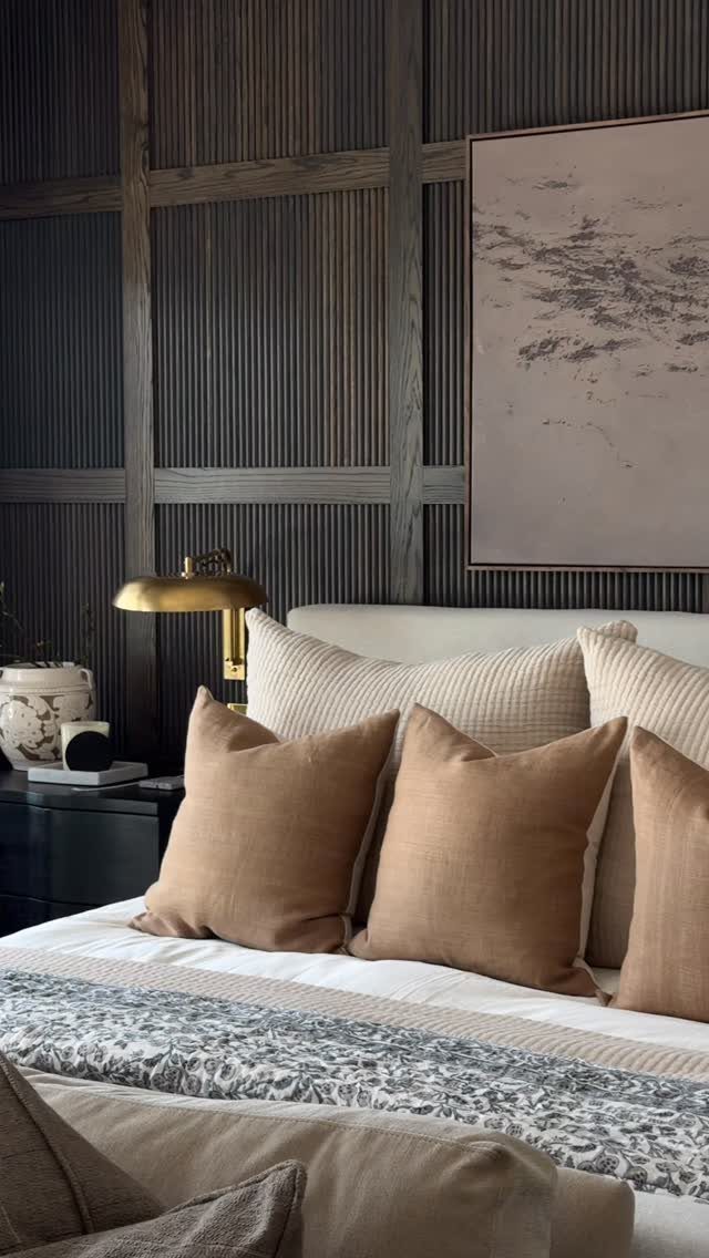 Bed with tan pillows against a dark ribbed wood paneled wall with artwork.