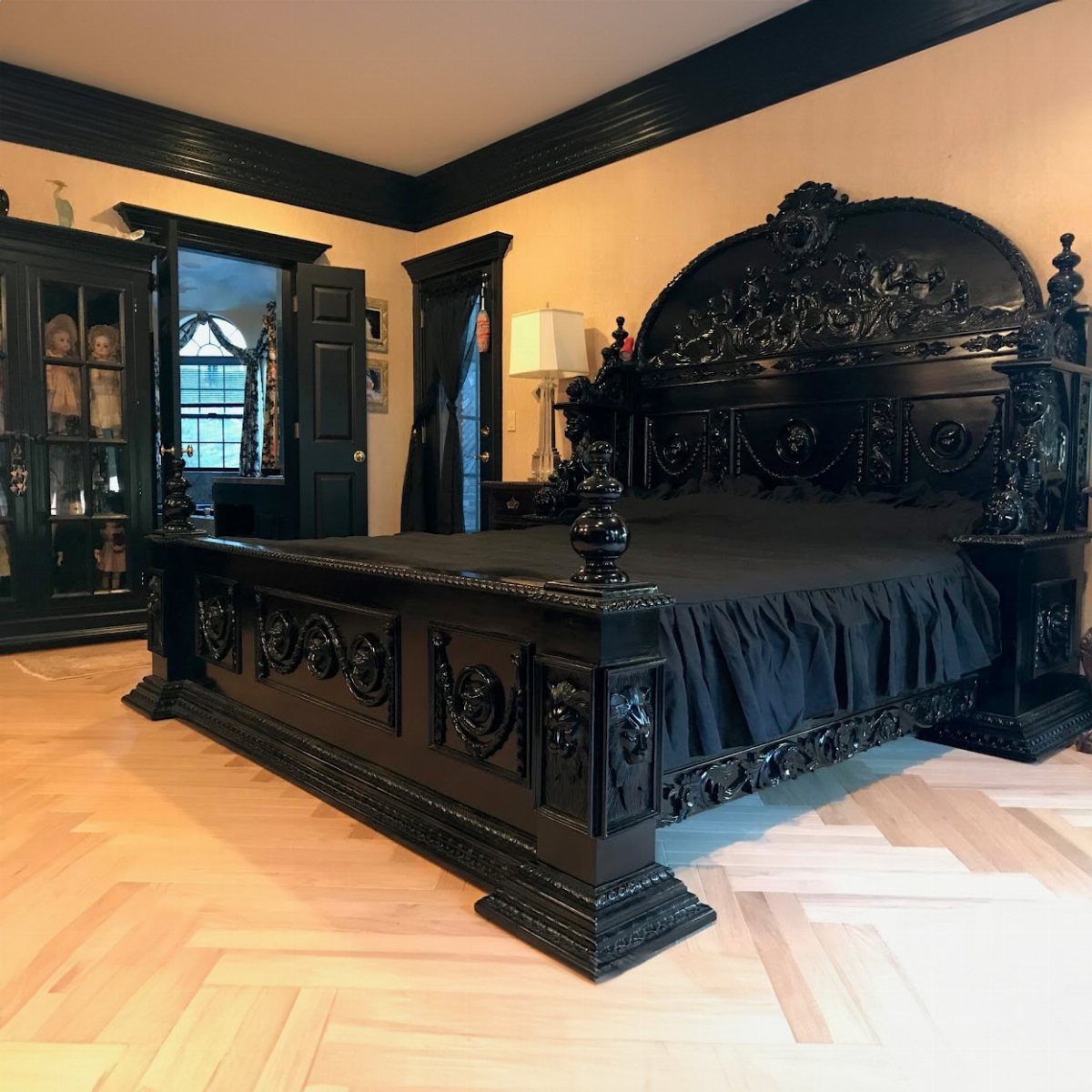 Ornate black carved wooden bed with black bedding on light herringbone wood floors.