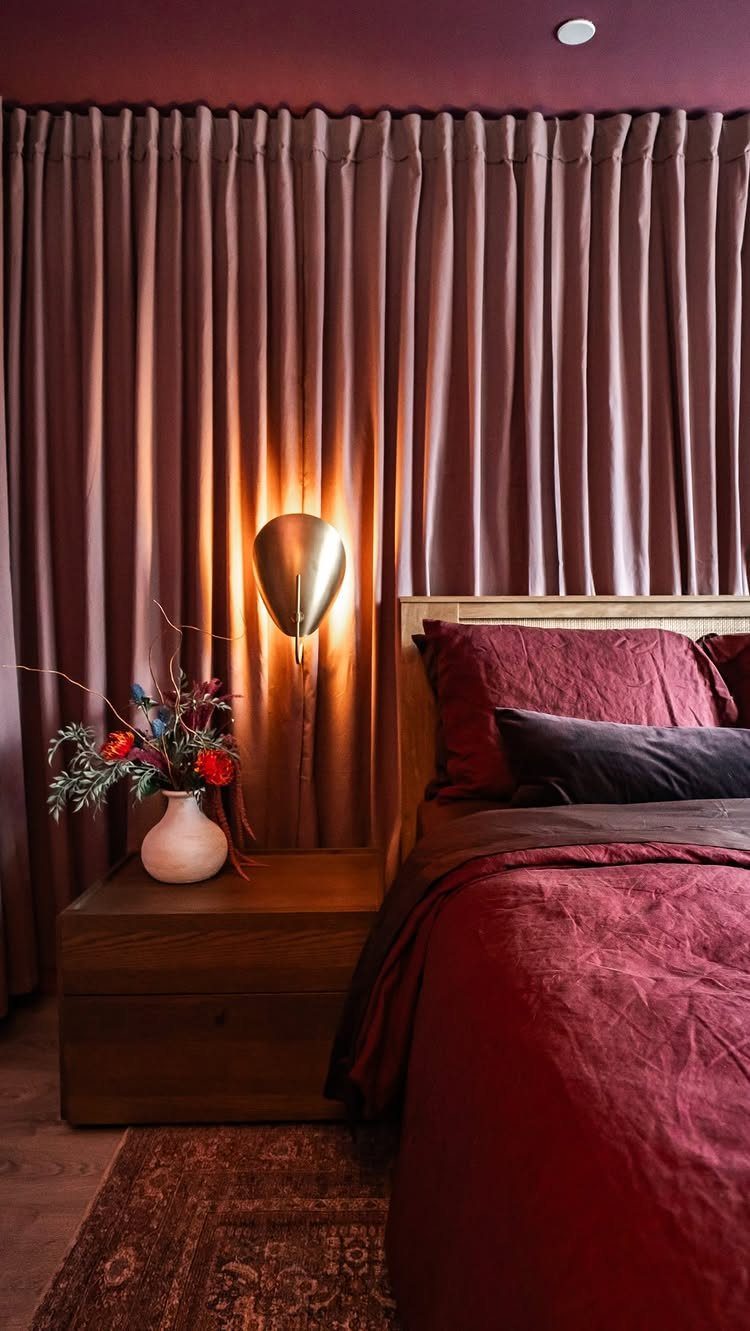 Mauve pleated curtains framing a bed with deep red linens and gold lighting.