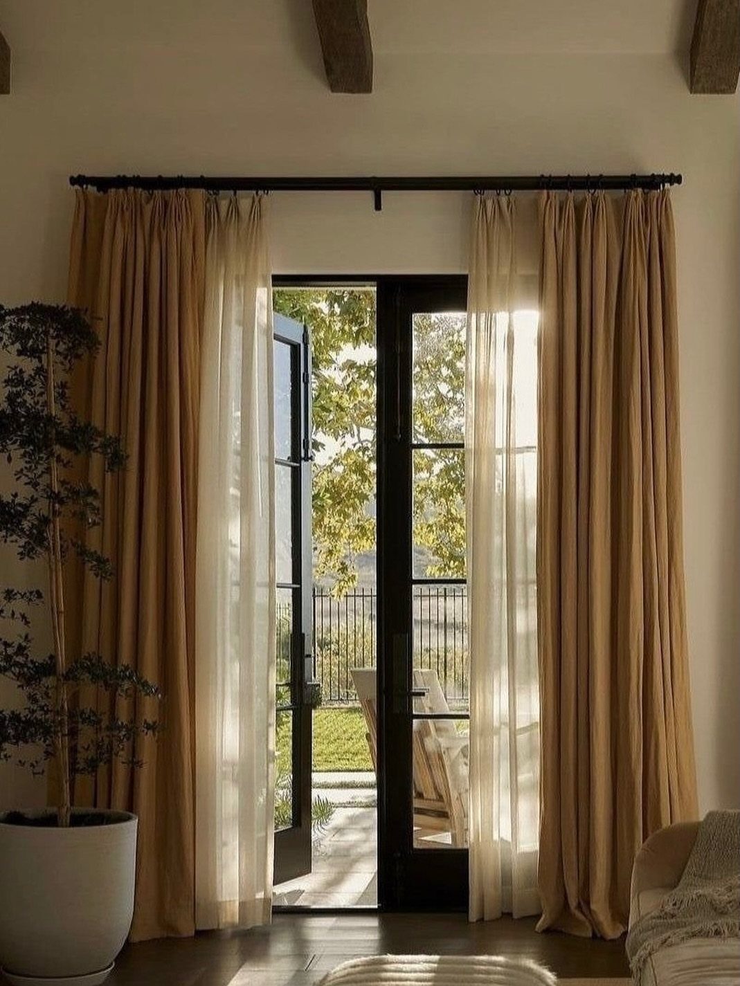 Layered tan blackout and white sheer curtains framing open black French doors.