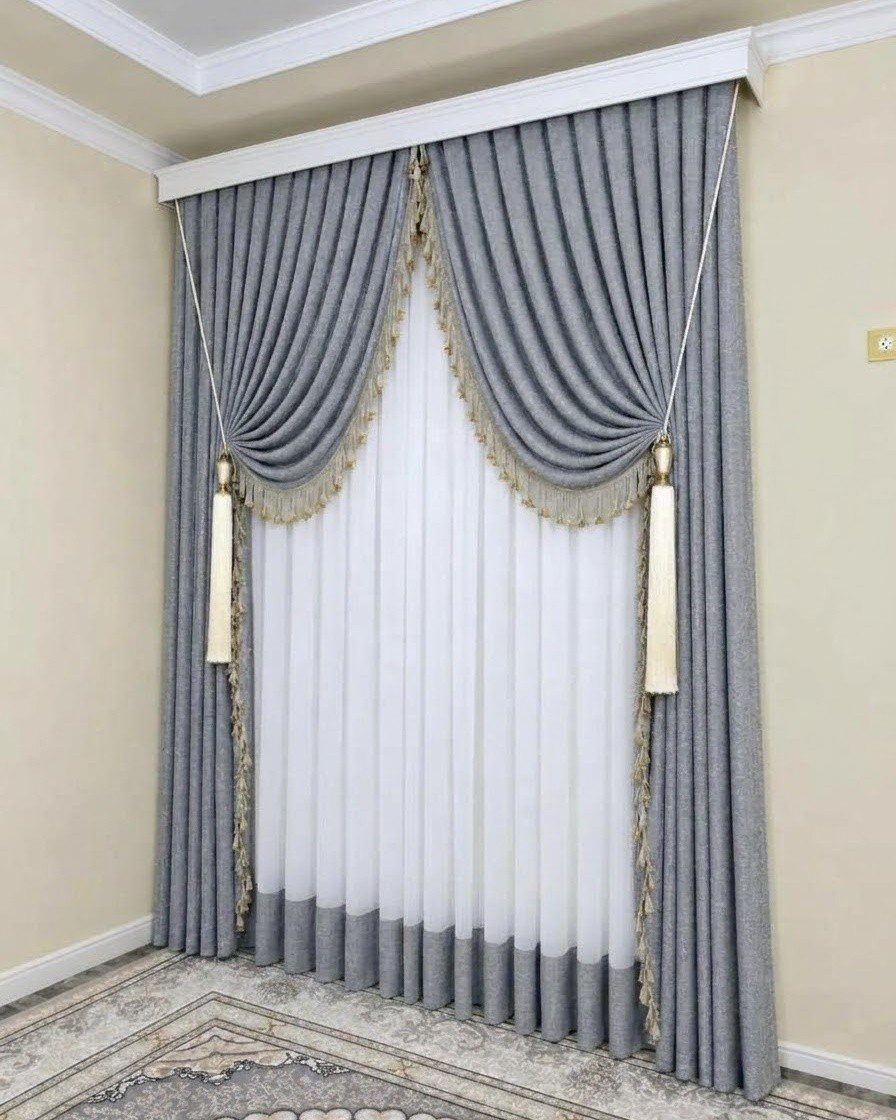 Grey draped curtains with gold tassels over white sheer curtains in a living room.