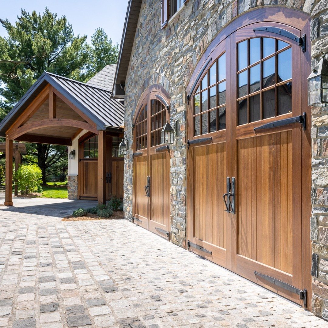 Arched wooden carriage-style garage doors set in a grey stone wall with cobblestone driveway.