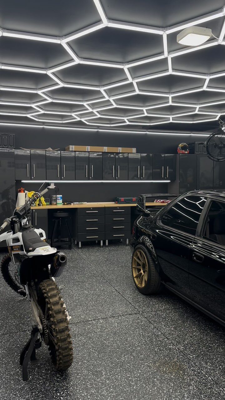 Modern garage workshop with hexagonal ceiling lights, black cabinets, a car and dirt bike.