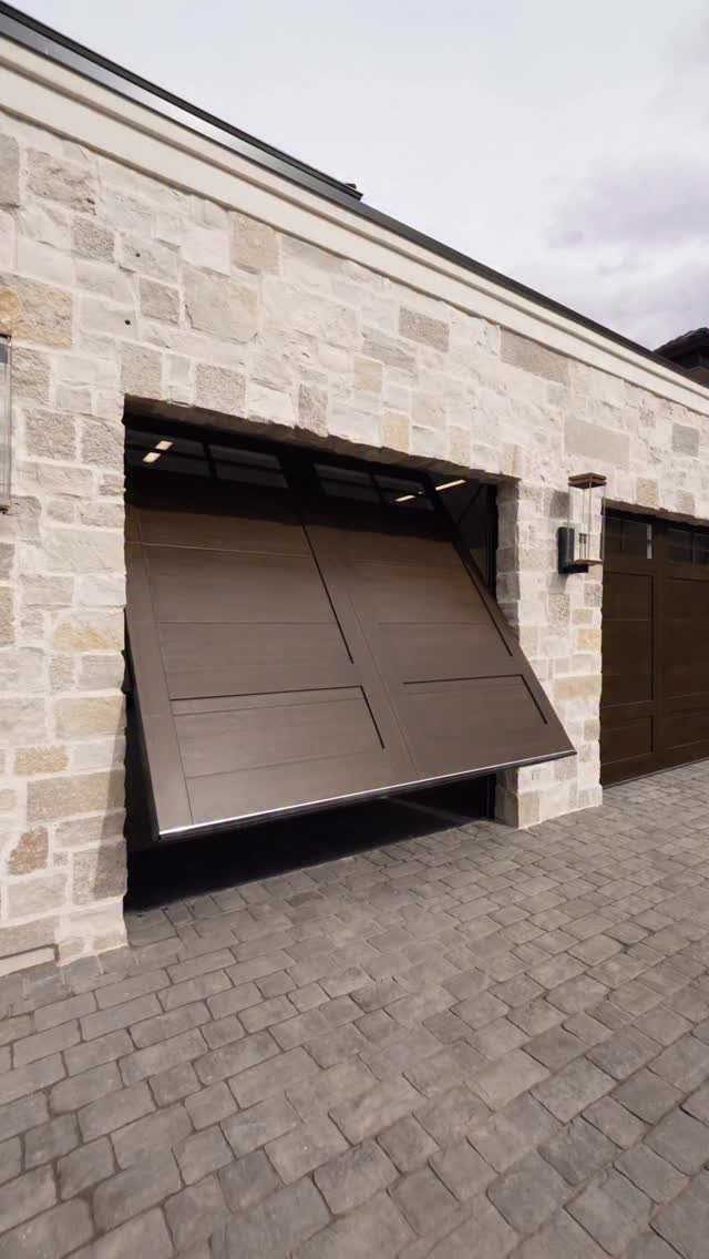 Brown tilt-up garage door opening outward from a light stone wall and paved driveway