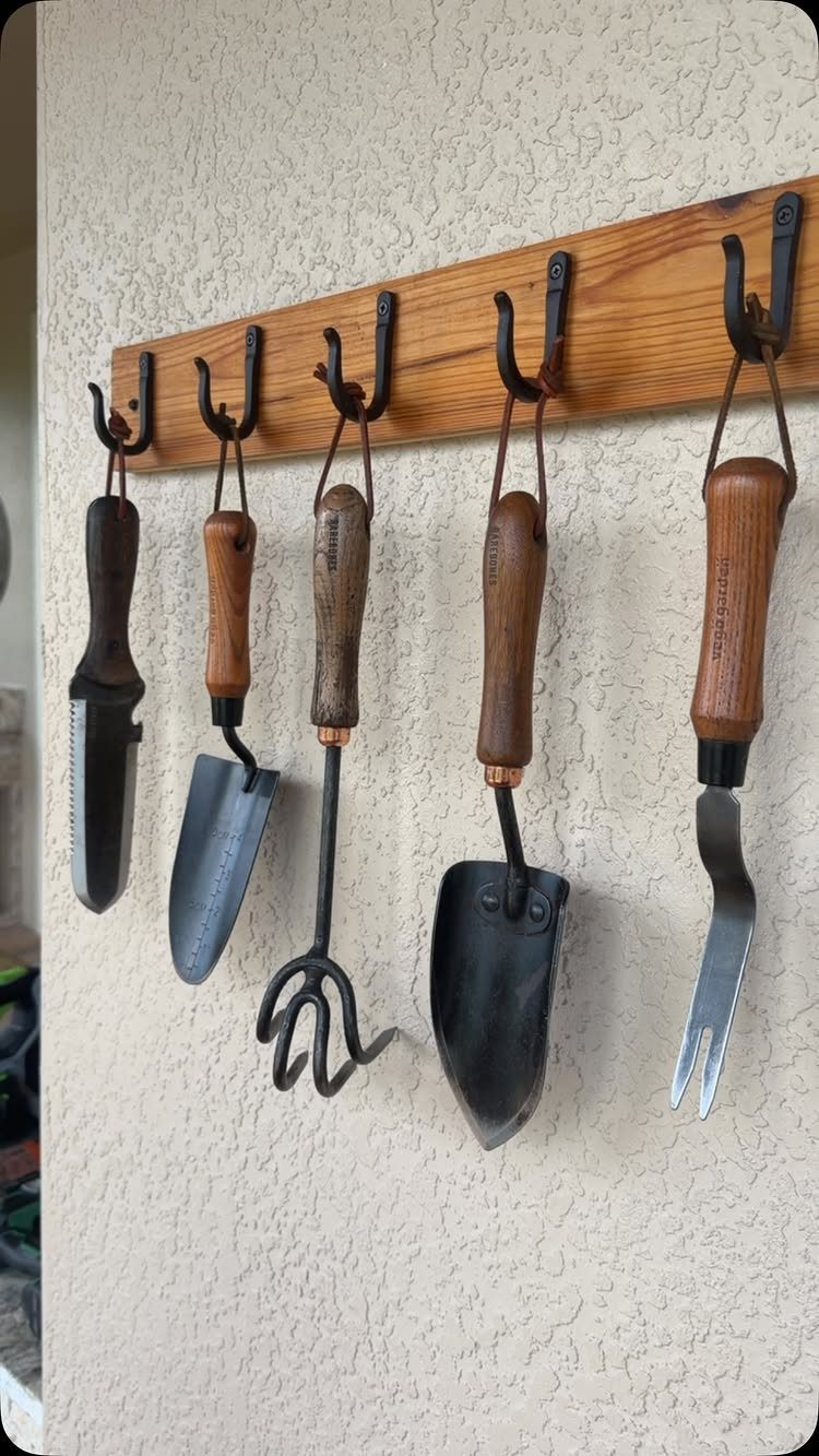 Hand gardening tools hanging from black metal hooks on a wooden wall rack.