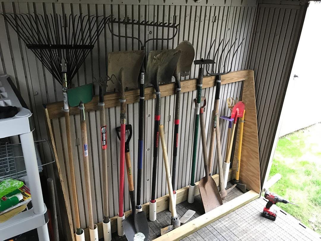 Garden rakes and shovels stored vertically in a wooden rack against a shed wall.