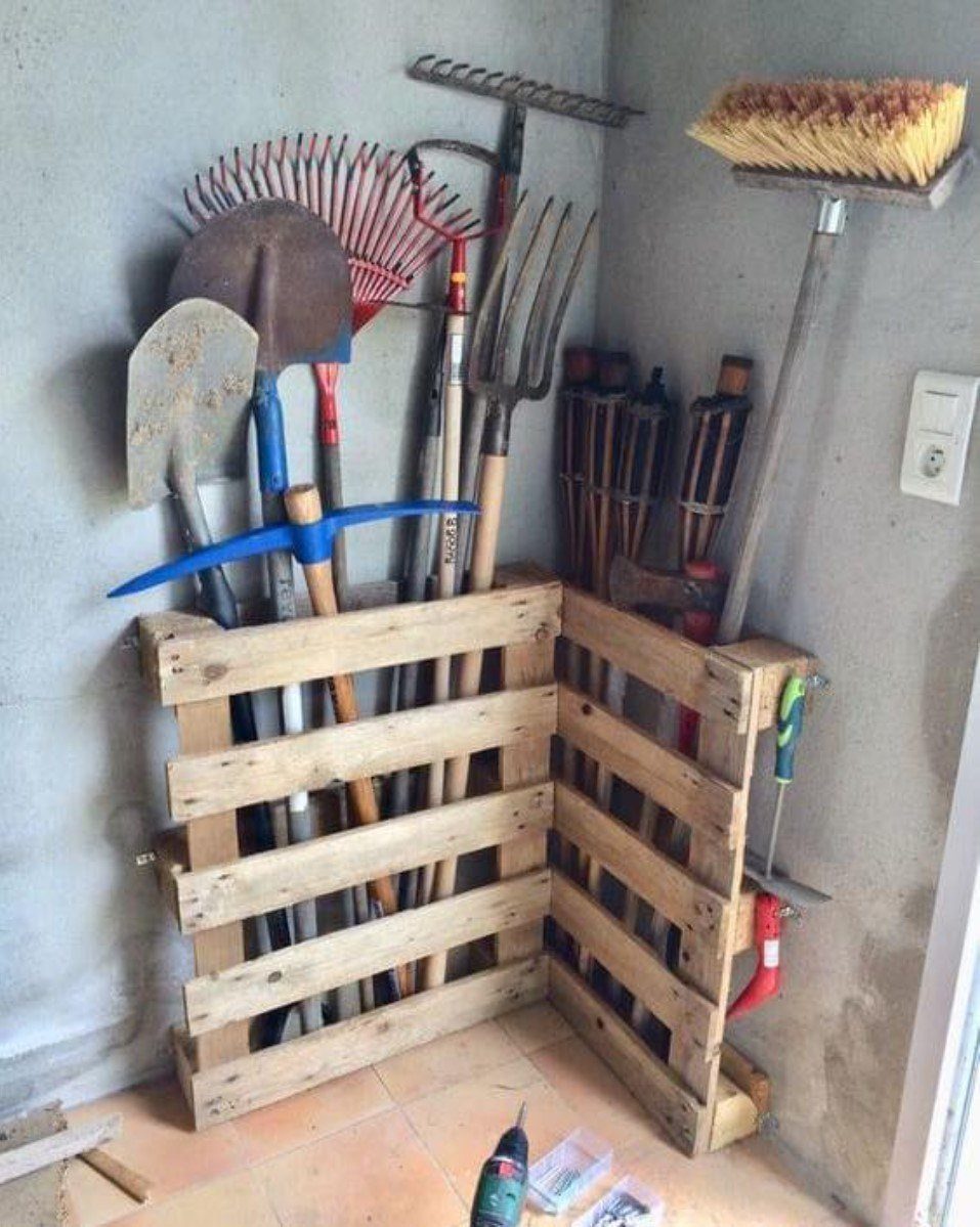 Wooden pallet racks holding garden shovels, rakes, and a broom in a room corner.
