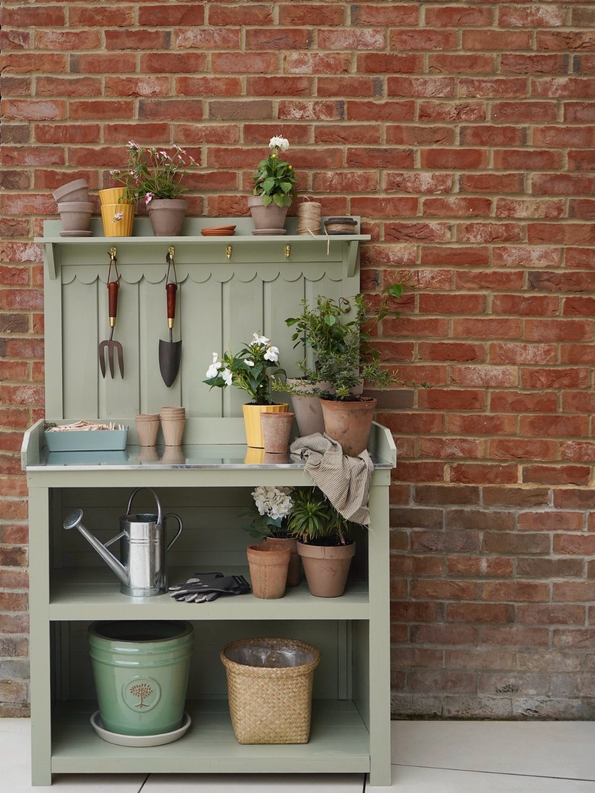 Green potting bench with hanging garden tools and potted plants against brick wall