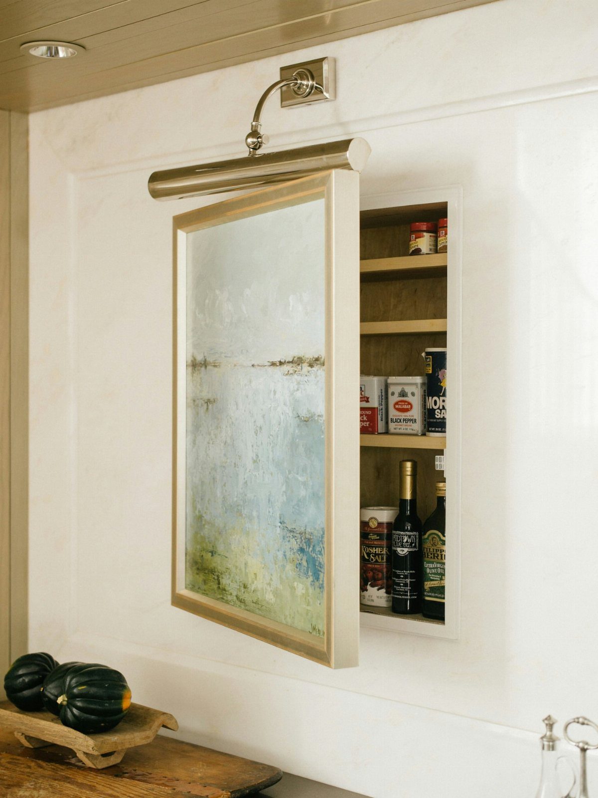 Framed painting swinging open to reveal hidden recessed wall shelves with wine and jars.