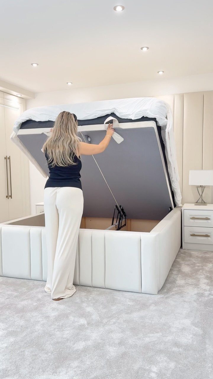 Woman lifting the mattress of a white upholstered storage bed in a modern bedroom.