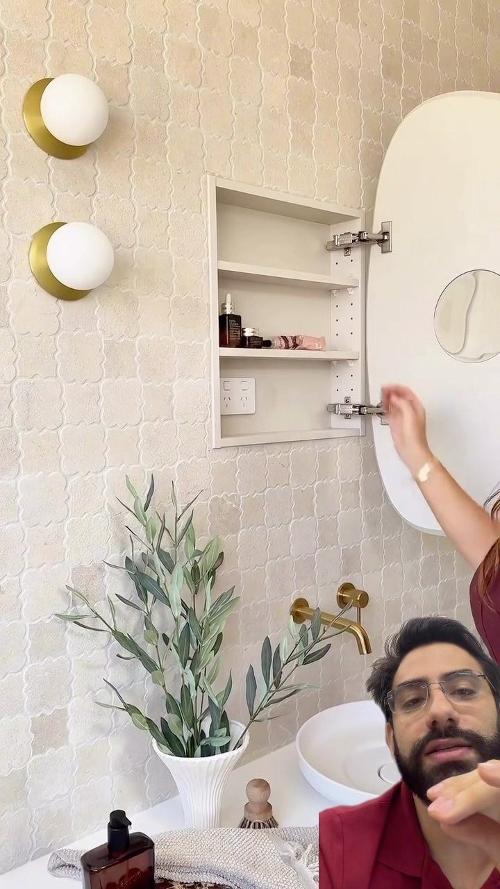 Oval mirror cabinet open to reveal white interior shelves on a tiled wall.