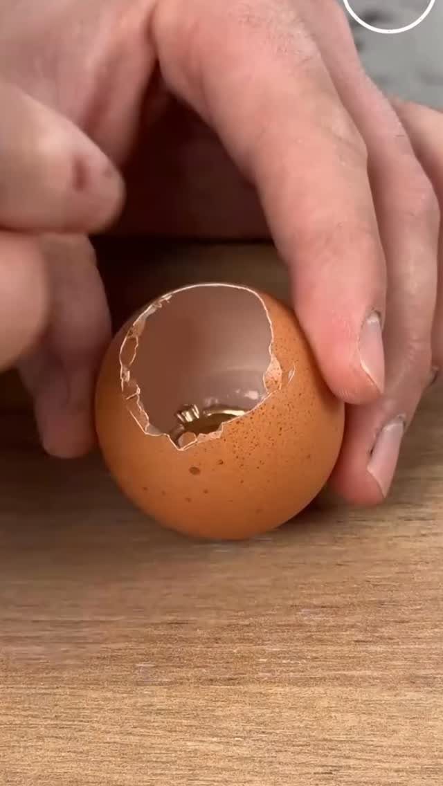 Brown eggshell held by hands with a gold ring hidden inside on wood.