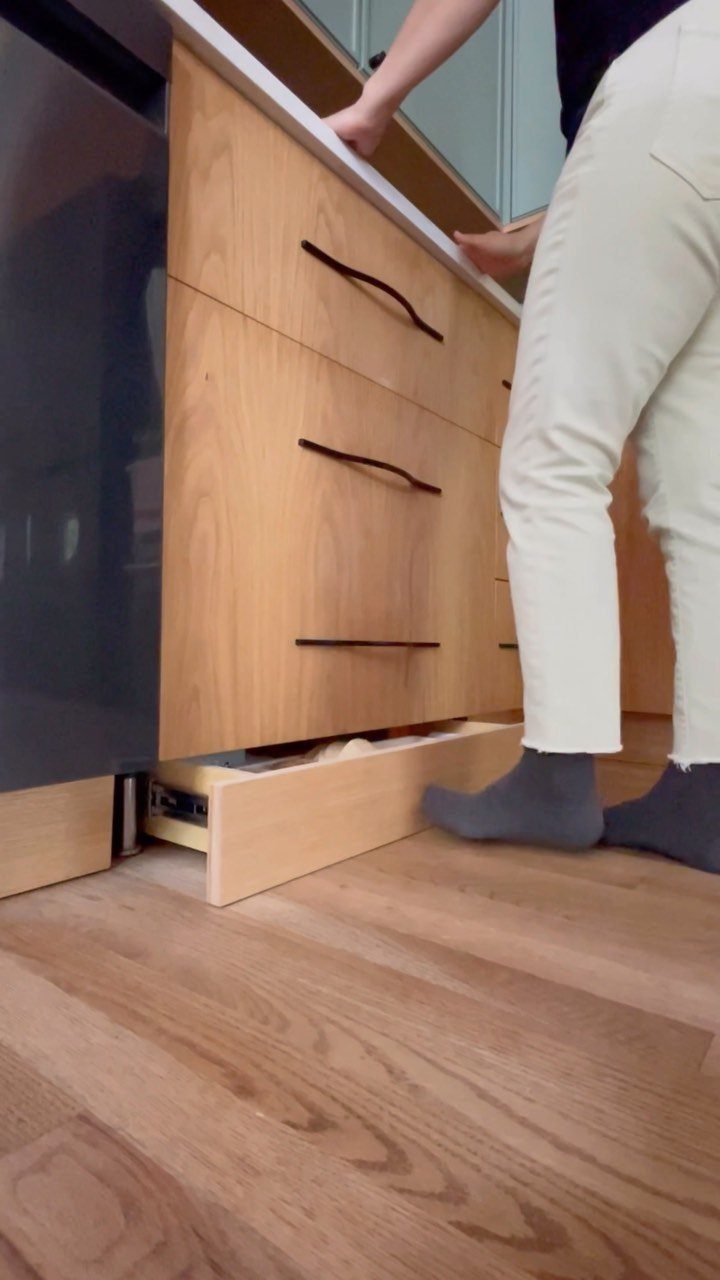 Toe-kick drawer pulled open at the base of light-wood kitchen cabinets.