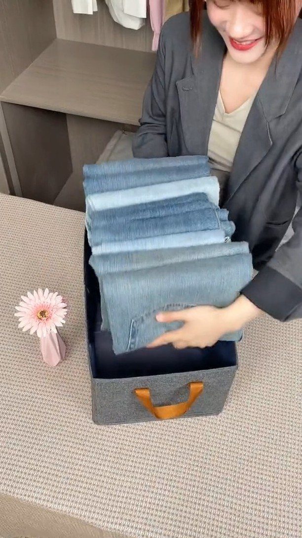 Woman placing a stack of folded blue jeans into a grey fabric storage bin.