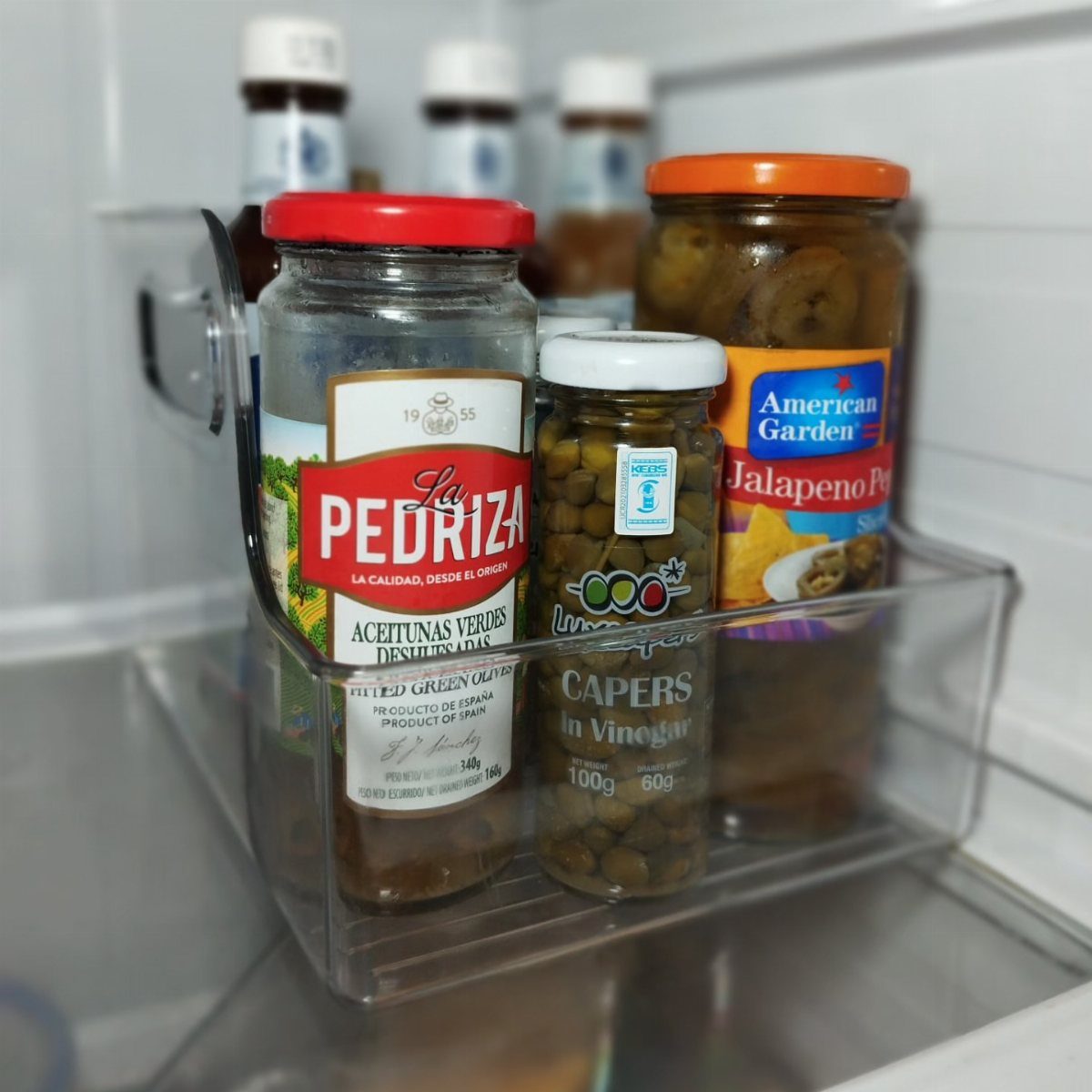 Clear plastic storage bin holding glass jars of condiments inside a white refrigerator.