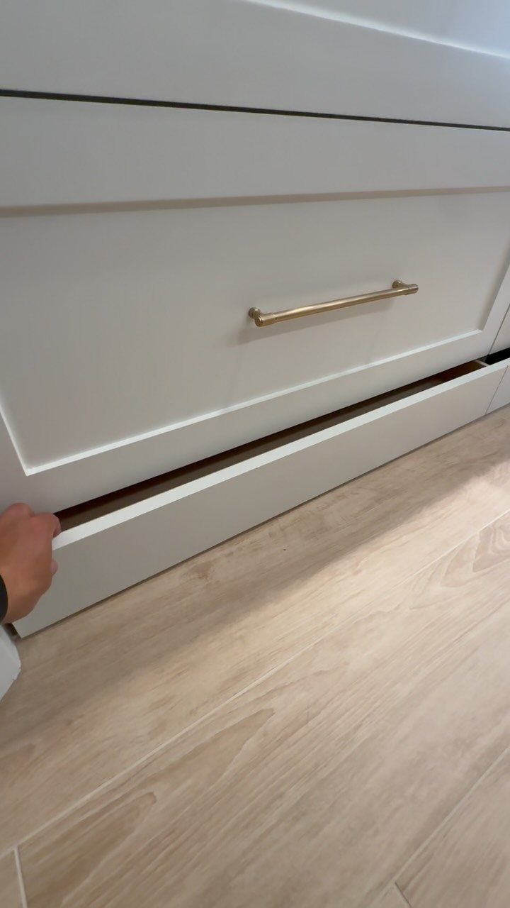Shallow white toe-kick drawer being opened beneath a kitchen cabinet on wood flooring.