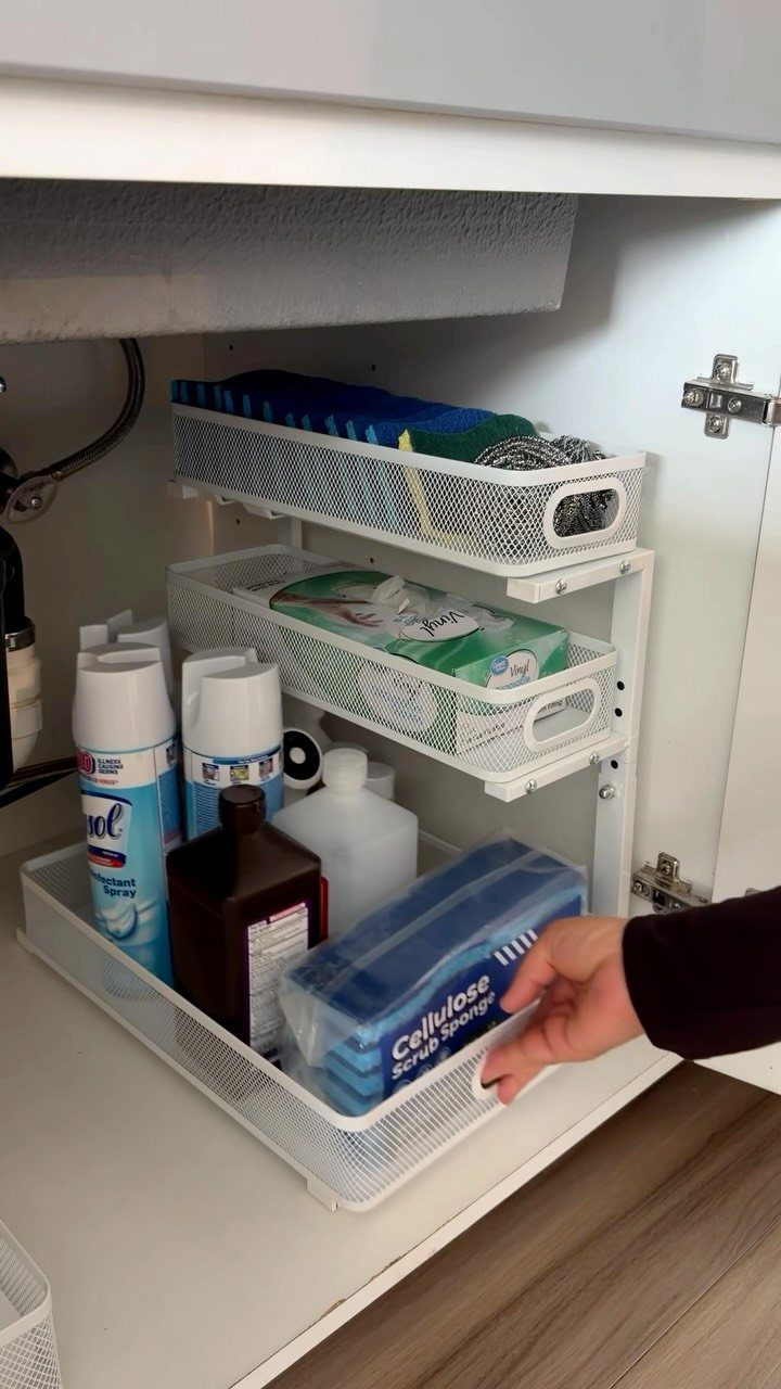 White metal pull-out organizer under a kitchen sink holding various cleaning supplies.