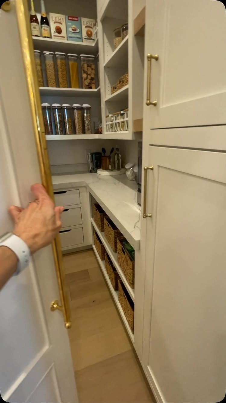 White kitchen pantry with gold handles, organized shelves, and woven storage baskets.