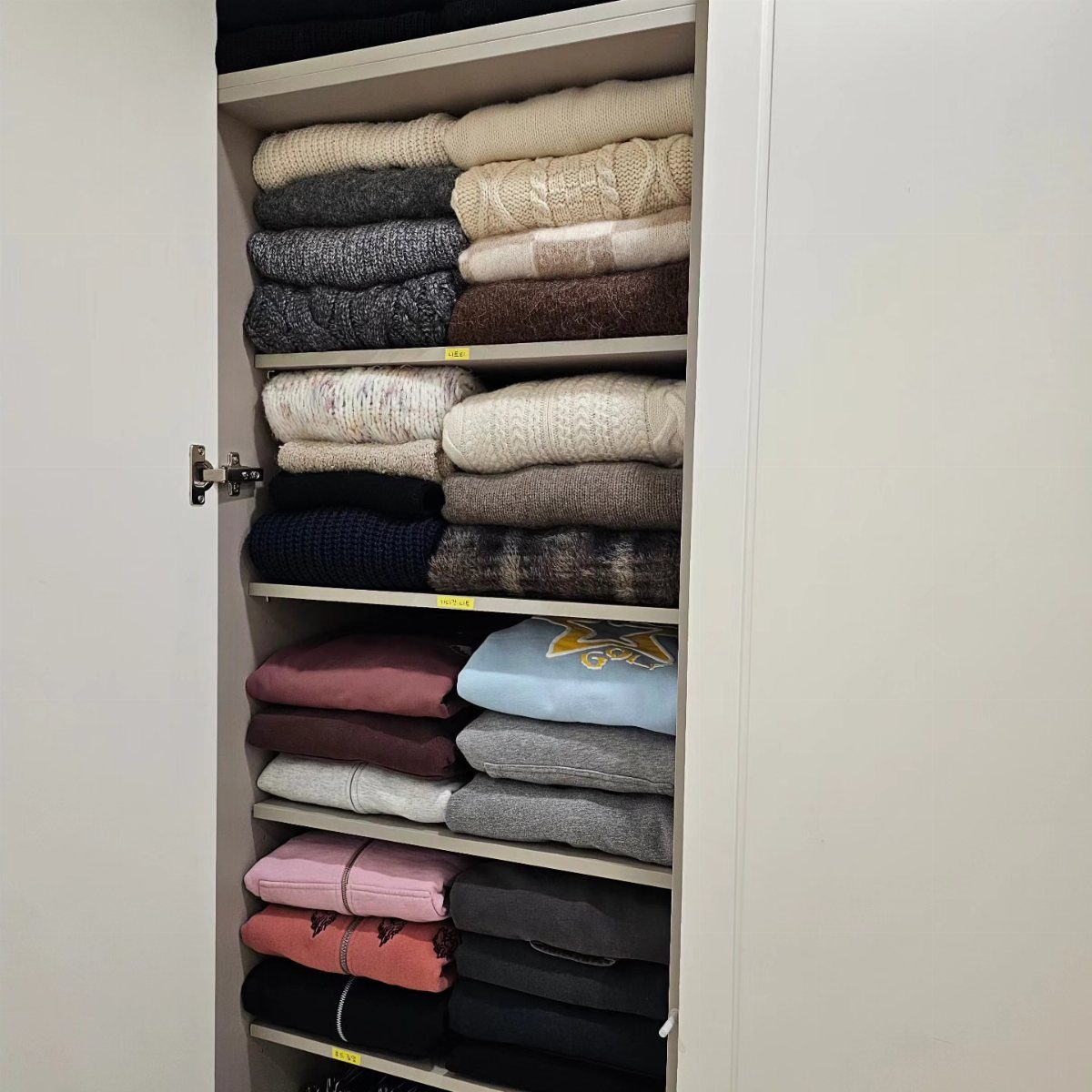 Folded sweaters in neutral and bright colors stacked on white closet shelves.