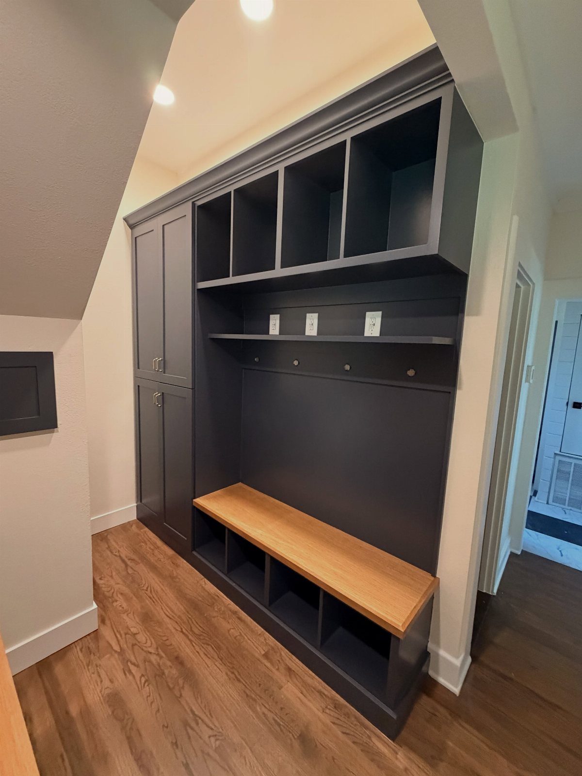 Dark grey built-in mudroom with wooden bench and cubbies under a slanted staircase.
