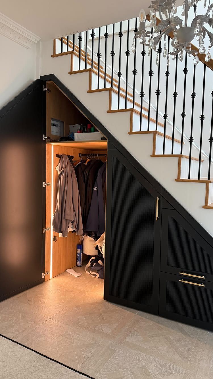 Black under-stairs coat closet with open door showing hanging outerwear and interior lighting.