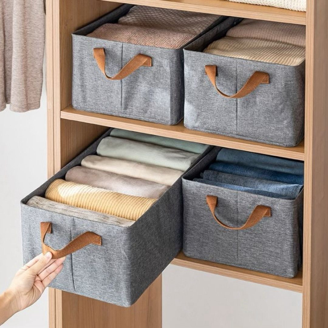 Grey fabric storage bins with brown handles holding folded clothes on wooden shelves.