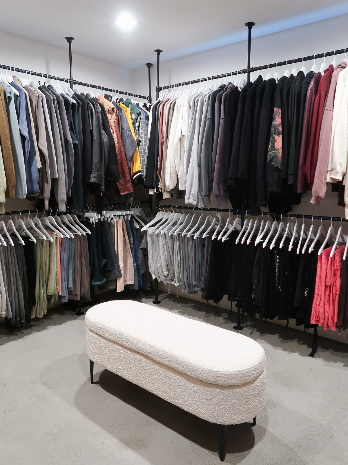 Two tiers of black hanging rods filled with clothes and a white ottoman.