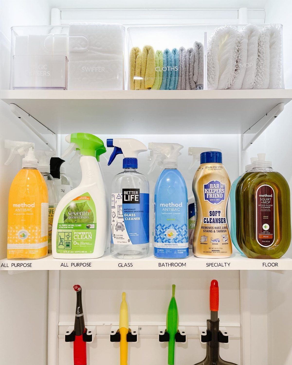 Organized cleaning closet with labeled spray bottles on white shelves and hanging tools.