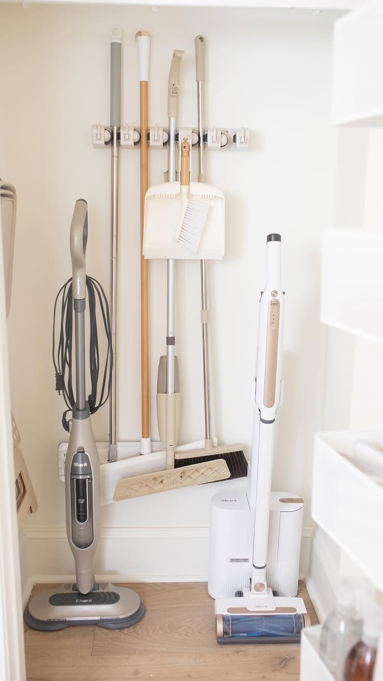 Organized cleaning closet with vacuums, brooms, and mops stored on white walls.