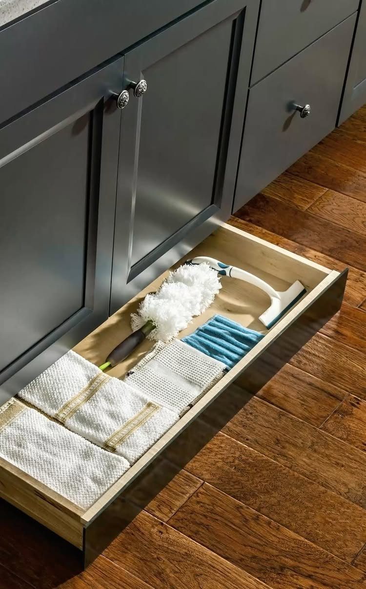 Shallow toe-kick drawer under grey kitchen cabinets containing cleaning cloths and a duster.