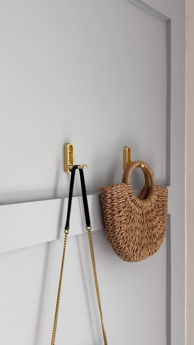 Gold wall hooks on a grey door holding a straw bag and chain purse.