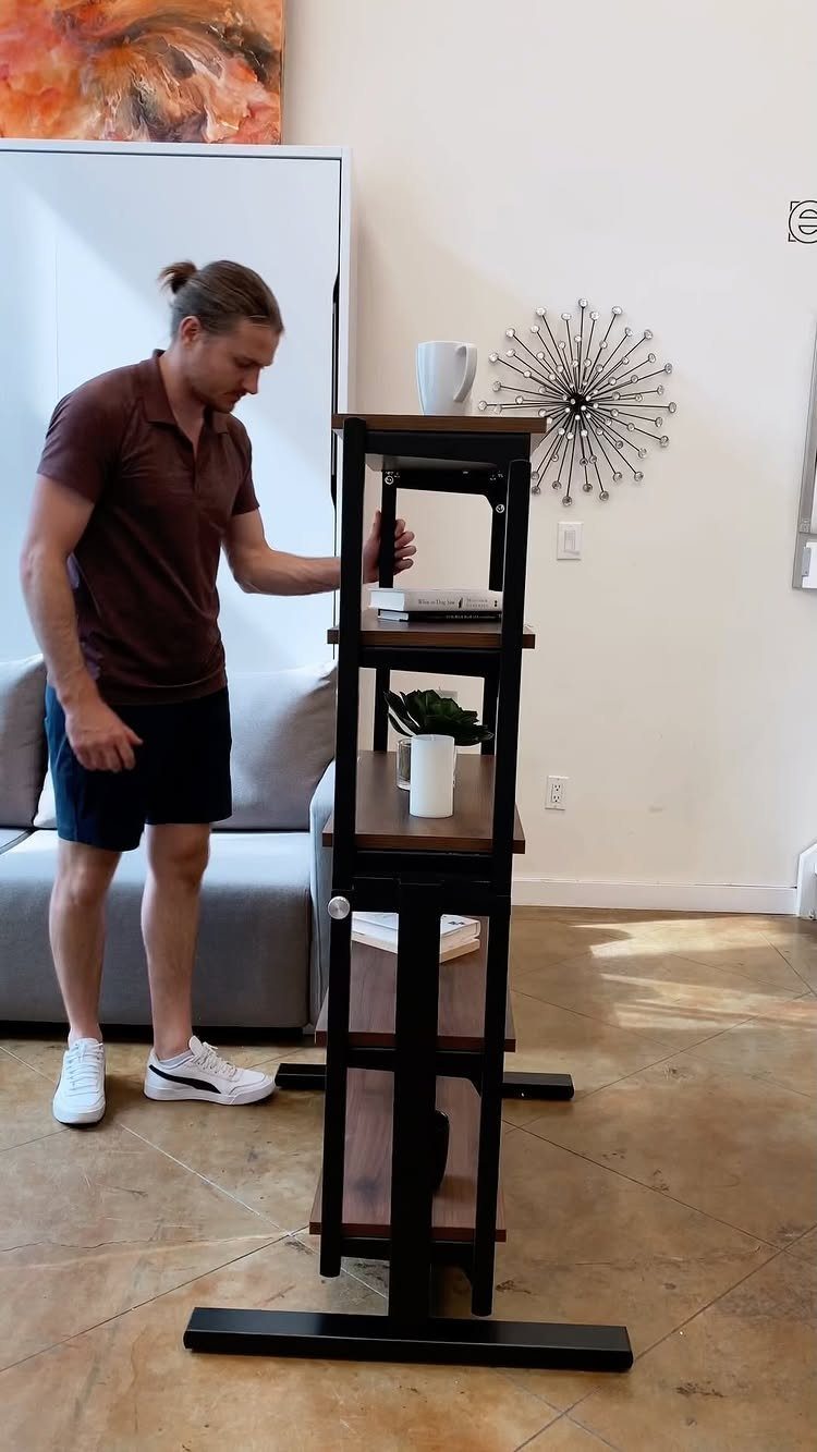 Man standing next to a tall narrow black metal shelf with wooden tiers