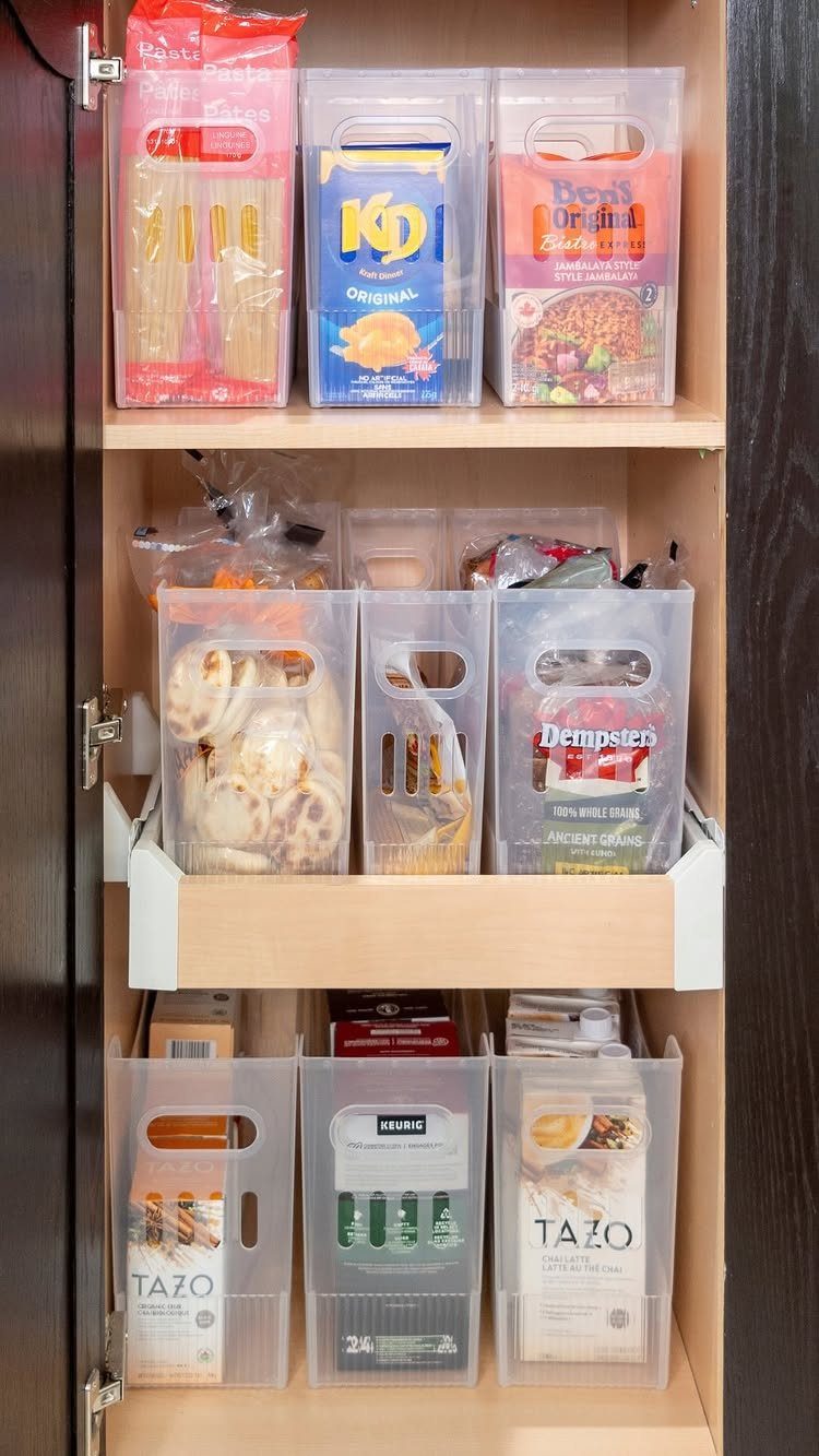 Clear plastic storage bins holding packaged snacks inside a wooden kitchen cabinet.