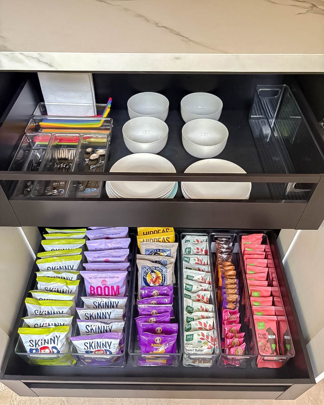 Clear plastic bins organizing colorful snack bags inside a deep dark kitchen drawer.