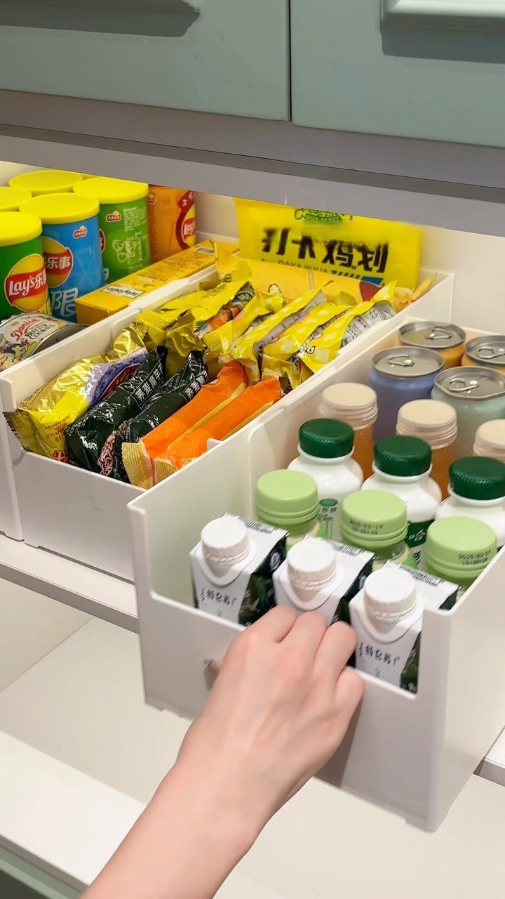 White plastic storage bins organizing packaged snacks and small bottles inside a cabinet.