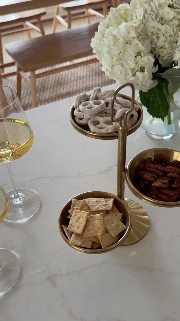 Gold three-tiered snack tray with pretzels, pecans, and crackers on a marble counter.