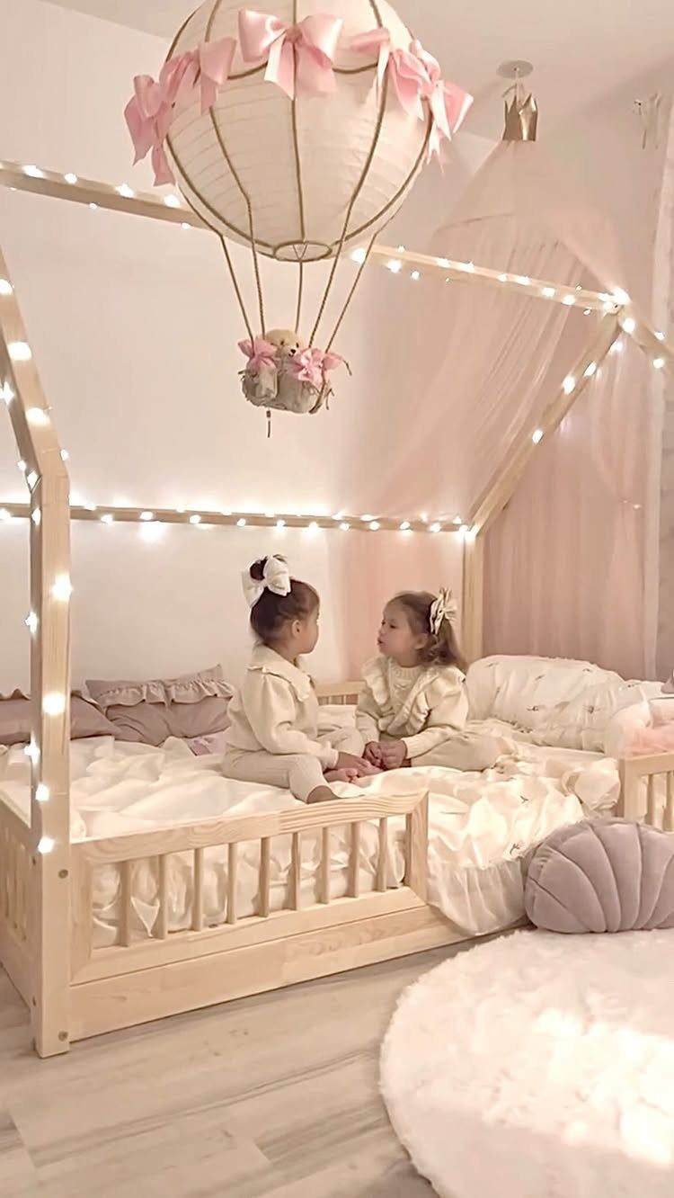 Two toddler girls sitting on a wooden house-frame bed with string lights.