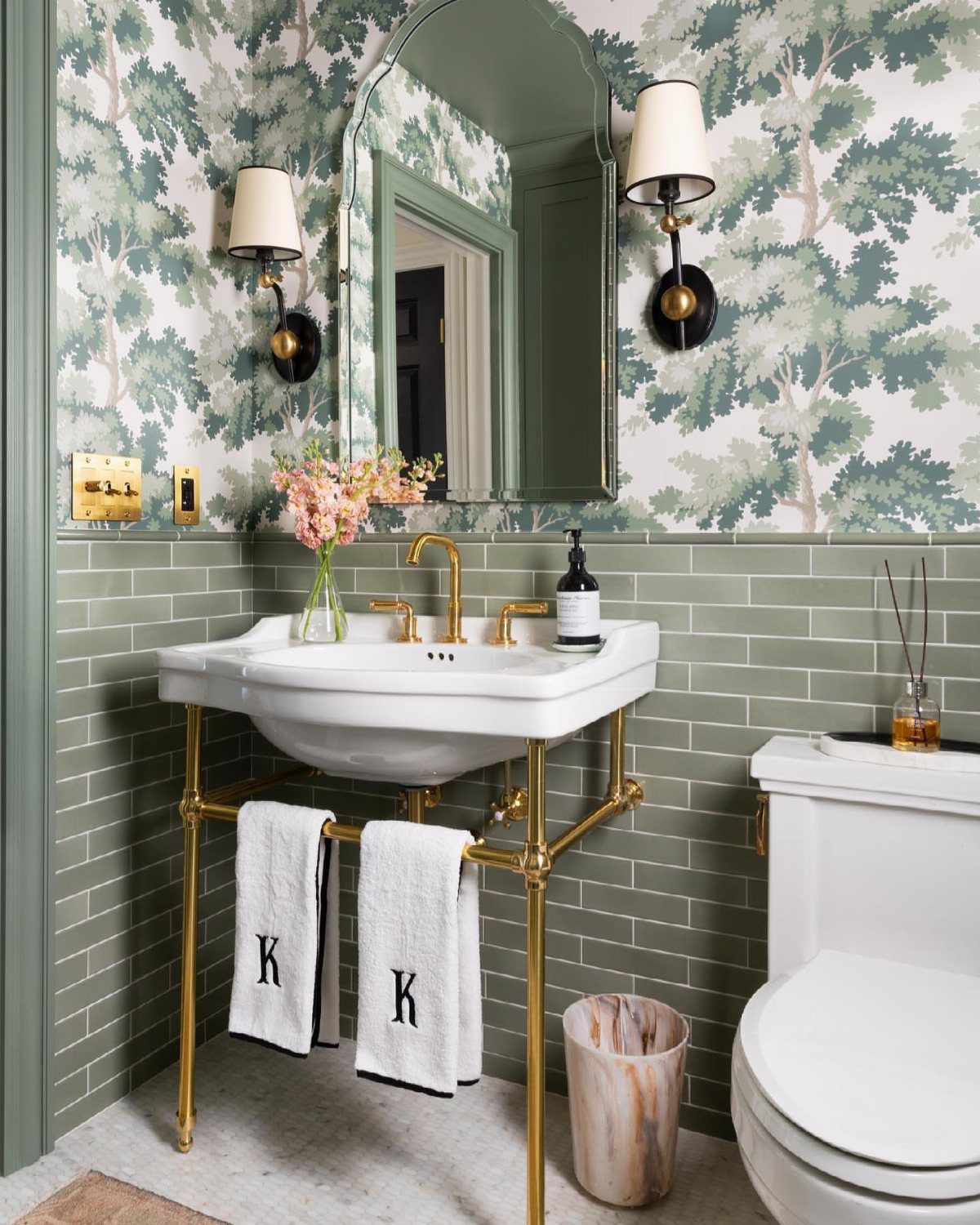 White console sink with gold legs against green tiles and forest wallpaper.