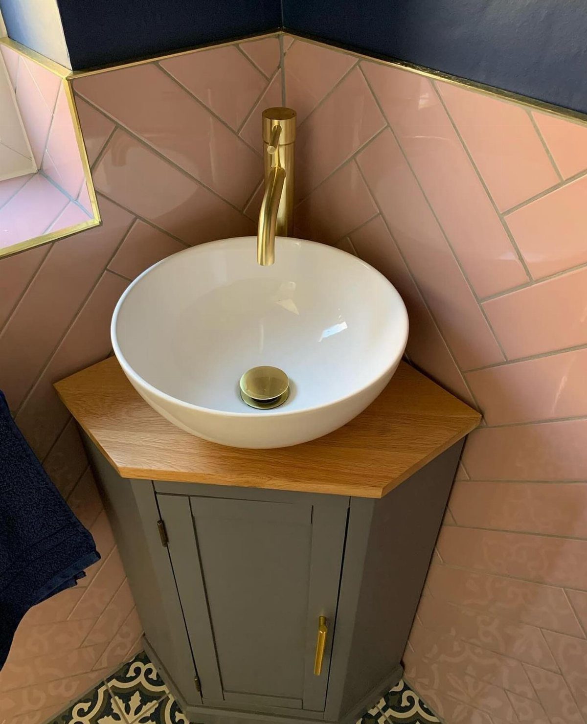 White vessel sink on a grey corner vanity with pink herringbone wall tiles.