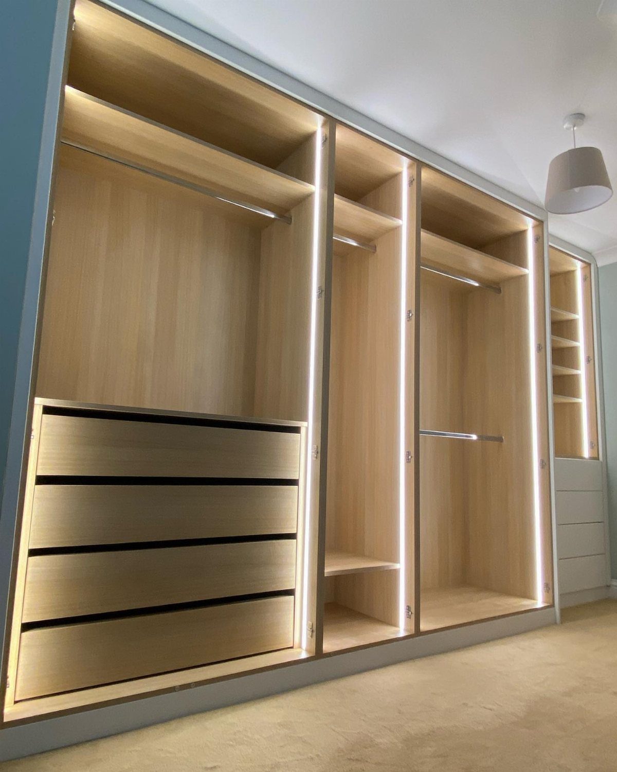 Built-in light-wood wardrobe with integrated vertical LED lighting strips and storage drawers.